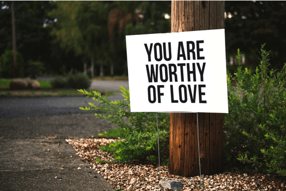 Self Love Is The Best Love, image of a white sign with the phrase you are worthy of love, sitting in front of a tree on a path