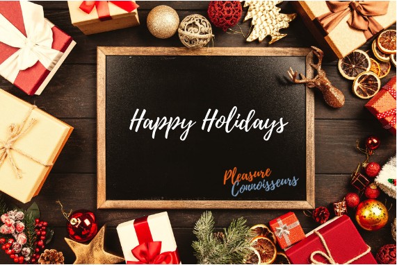 Happy holidays, chalboard with the words happy holidays in white and the Pleasure Connoisseurs logo in orange and blue, the board is surrounded by gifts wrapped in red and gold.